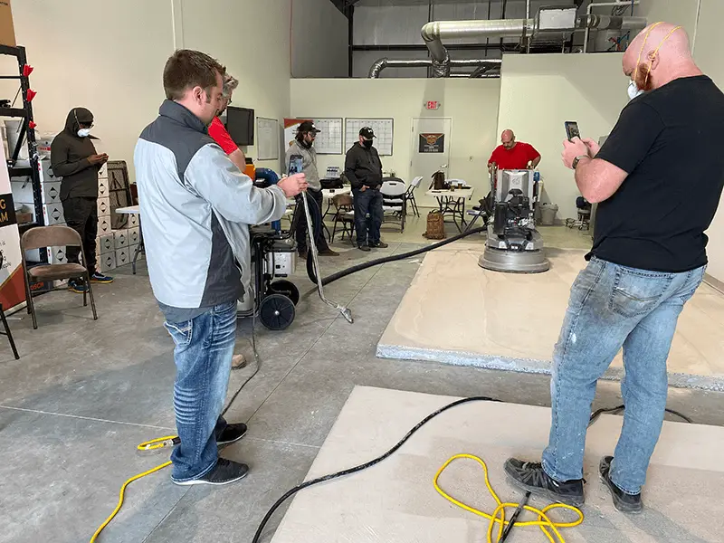 A group of people in a workshop setting is gathered around a concrete grinding machine. Some are observing, while others appear to be taking notes or photos. The room has industrial equipment, and various tools are lying on the floor.