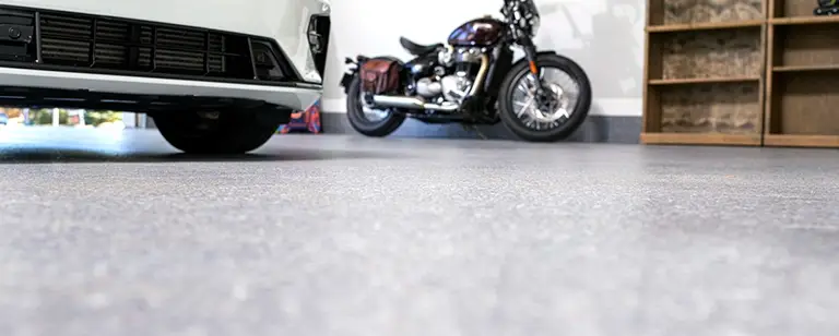 A garage with a white car parked on the left and a motorcycle in the background. The floor is covered with a smooth, polished concrete surface. Shelving units are visible on the right side of the image with items stored on them.