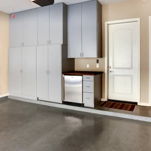 A clean and organized garage interior with gray cabinets and a mini refrigerator under a wooden countertop. The polished concrete garage floor reflects light, potentially minimizing condensation causes. A white door with a red and brown striped mat is seen on the right side of the image.