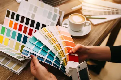 A person is holding multiple color swatches or sample books, displaying a variety of garage floor colors. There's a white coffee cup on a saucer, a pen, and a smartphone on the wooden table, with a laptop partially visible in the background.