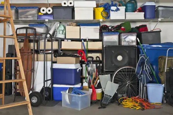 A cluttered garage filled with various storage bins, shelves, and miscellaneous items, including a ladder, cooler, bicycle wheel, garden tools, and electrical cords. Cabinets and shelves hold boxes, plastic containers, and camping gear in this disorganized storage system.