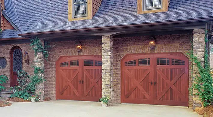 A two-story house with a brick exterior and a steep, shingled roof. The house features two large wooden garage doors, each with a pair of lantern-style light fixtures above. Climbing plants are growing around the stone columns between the garage doors, enhancing its Garage Kings appeal.