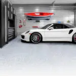 A white sports car is parked in a sleek, modern garage. The wall features black tool cabinets, a red kayak mounted on the wall, and various tools and equipment arranged neatly. The floor is clean and smooth, and a partially open garage door is seen on the right.