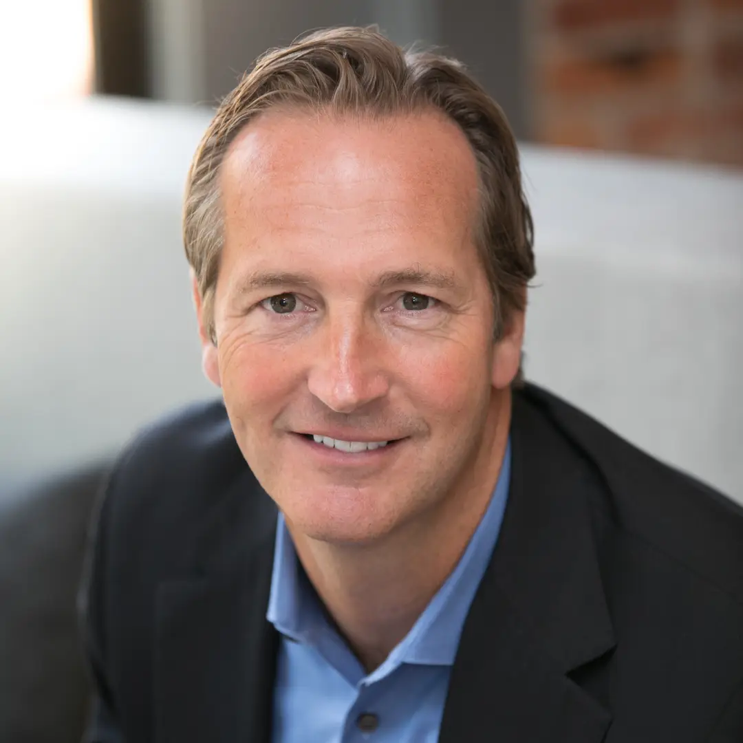 A middle-aged man with light skin, short brown hair, and blue eyes is smiling at the camera. He is wearing a blue dress shirt and a dark blazer. The background is softly lit with a mix of light and dark areas, giving a professional setting feel.