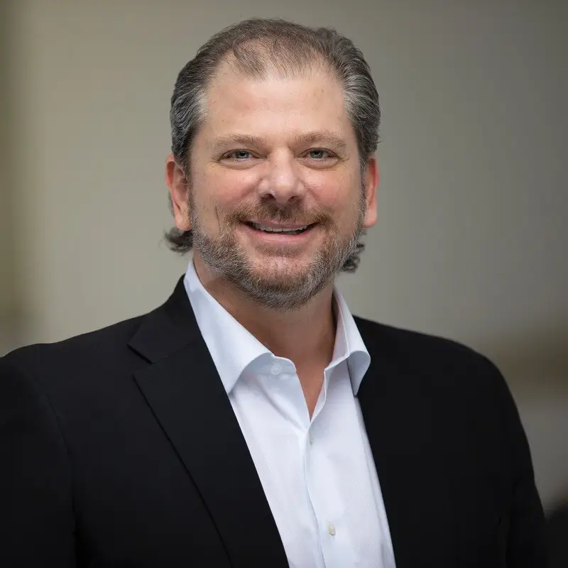 A man with short, graying hair and a beard smiles at the camera. He is wearing a white dress shirt and a dark blazer. The background is blurred, drawing focus to the man.