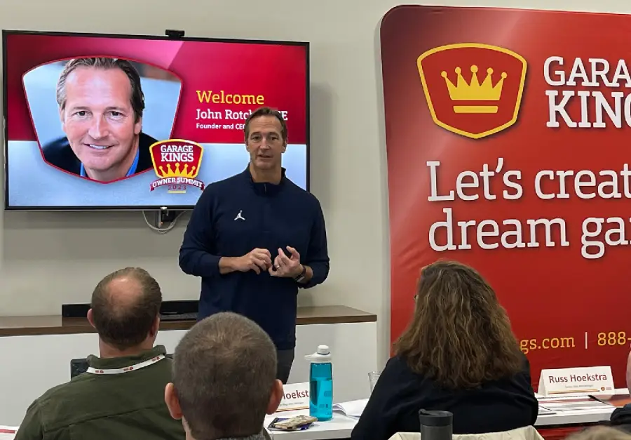 A man stands in front of a group, speaking. Behind him, a screen displays "Welcome John Rotche, CFE," with a photo of the same man and a Garage Kings logo. A red banner with "Garage Kings" and text "Let's create a dream garage" is to the right. Seated audience members are visible.