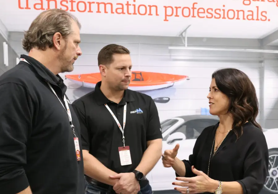 Three individuals are engaged in a discussion in front of a wall with a banner stating, "transformation professionals." The man on the left wears a black shirt, the man in the middle also wears black, and the woman on the right is gesturing while speaking.