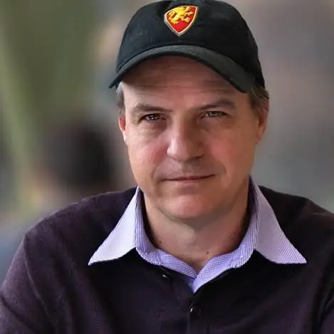 A middle-aged person with fair skin wearing a black baseball cap with a red and gold emblem. They have short, light brown hair and are dressed in a dark sweater over a light blue collared shirt. They are looking directly at the camera with a neutral expression. The background is softly blurred.