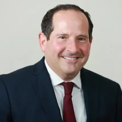 A man with short dark hair is smiling at the camera. He is wearing a dark suit, white dress shirt, and a red tie. The background is plain and light-colored.