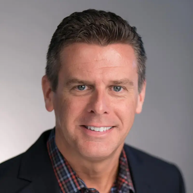 A man with short, dark hair smiles at the camera. He is wearing a checkered shirt under a dark blazer. The background is a gradient of light gray, transitioning from lighter on the left to darker on the right.