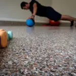 A person in an exercise room is performing a plank with one hand on a blue medicine ball and the other on the floor. In the foreground, there are different colored weights and a kettlebell placed on a speckled floor.