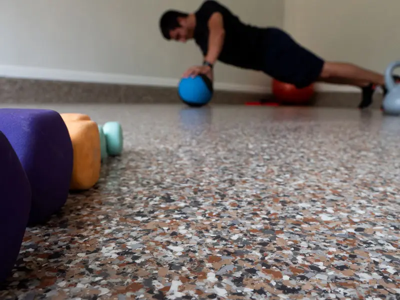 A person is doing push-ups on a medicine ball in a gym. The focus is on a set of colorful dumbbells in the foreground on a speckled floor, while the person in exercise clothing is blurred in the background.