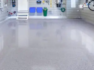 A clean, organized garage with gray walls and a shiny, epoxy-coated floor. Shelves on the walls hold various tools, two blue storage bins, a green trash bin, and a coiled garden hose. A step leads to a closed door in the background.