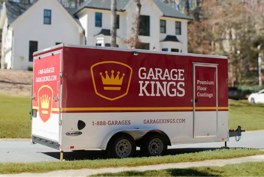 A red and white trailer with the "Garage Kings" logo and crown emblem is parked on a residential street, showcasing premium floor coatings. Displaying a phone number and website for those eager to get started with this renowned franchise, houses are visible in the background.