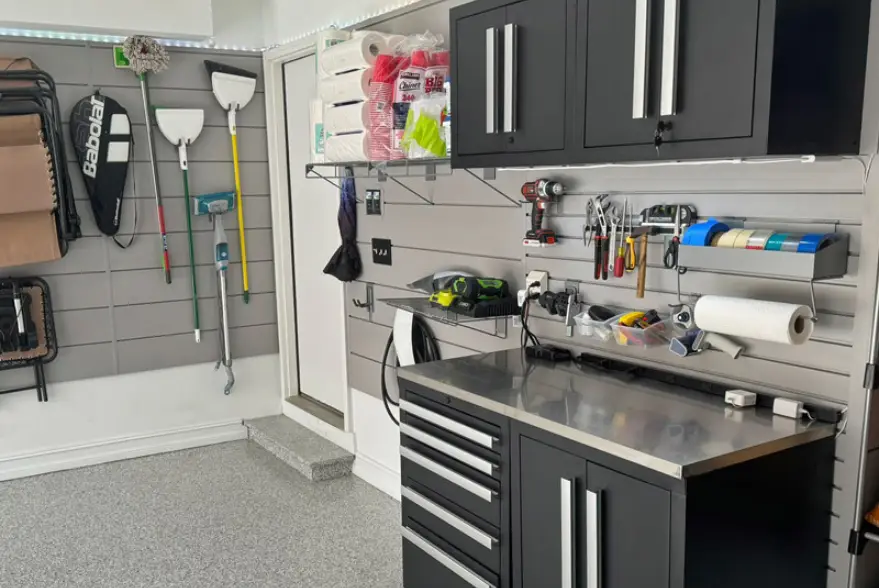 A neatly organized garage invites you to get started on any project. It features black cabinets, a stainless steel workbench, and an array of items like paddles and brooms mounted on the walls. The floor boasts a gray speckled pattern—perfect for setting up your own personal workshop franchise.