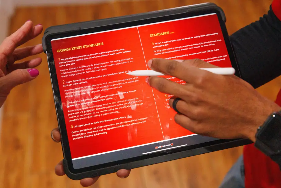 Two people interact with a tablet displaying text on a red background. One holds the tablet while the other points at the screen with a stylus. The document appears to outline several procedural standards, perfect for getting started on your new garage franchise journey.