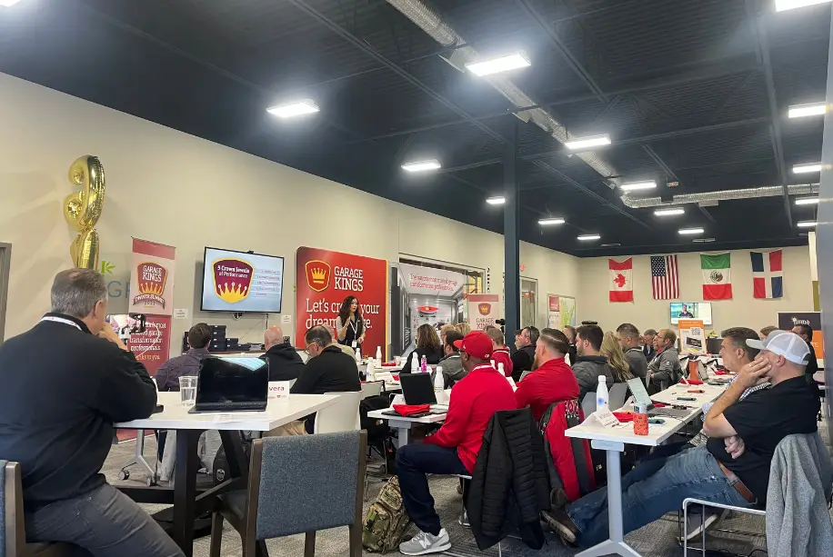 A group of people sit at tables in a conference room, eager to get started with a presentation on garage franchise opportunities. Flags from different countries hang on the wall, while posters and a large screen command attention at the front of the room.