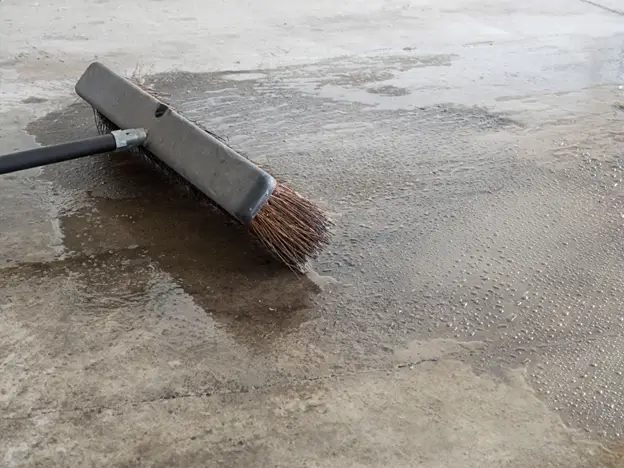 A broom with a long handle efficiently sweeps water from the garage floor's concrete surface. The ground is partially wet and drying, while the dark brown bristles, made from natural fibers, ensure excellent garage floor maintenance.